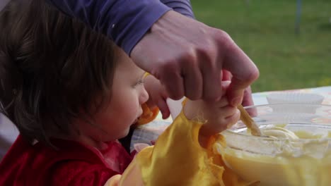 Close-up-of-small-child-and-mother,-helping-spoon-cake-mixture-into-cases-on-a-patio-table-during-the-daytime