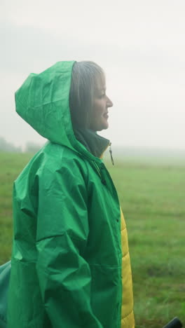 side view of woman in green raincoat carrying mint-colored bag and umbrella, walking on a grassy field under misty sky, looking into the distance, surrounded by open greenery and cloudy sky