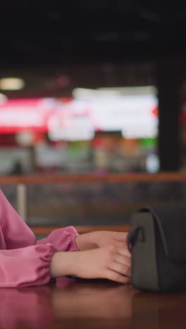 dama blanca en vestido rosa se sienta en un centro comercial con su bolsa en una mesa de madera, abriendo su portátil para trabajar, el fondo está borroso con la gente moviéndose, con un efecto de luz bokeh