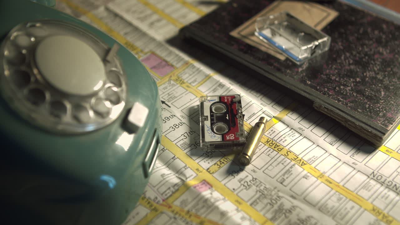 Close up reveals a mini cassette recorder, spent bullet shell, and vintage phone on a city map. High-detail crime scene evidence