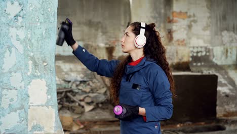 urban street artist in headphones is listening to music and painting on the wall in old empty house. pretty girl is using aerosol spray paint to create beautiful image.