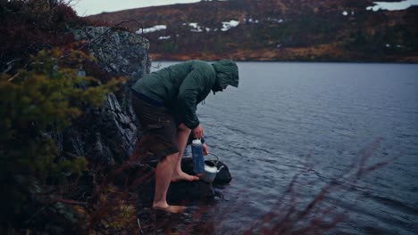 man wearing a hoody sweatshirt filling a bottle tumbler in a lake. static shot