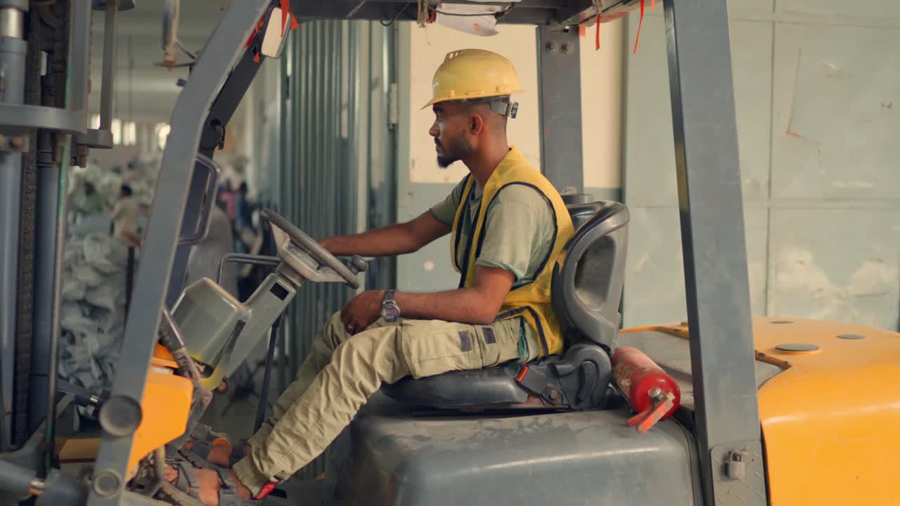 Ripresa orbitale dinamica a mano di un operaio in forma che indossa un gilet di sicurezza giallo e un casco mentre utilizza un carrello elevatore grigio e giallo all'interno di una fabbrica di tende molto trafficata a Sialkot
