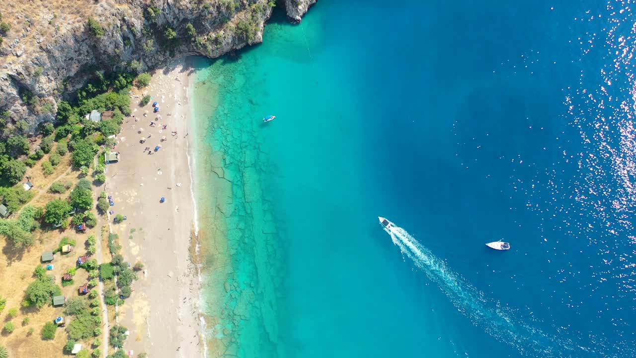 aerial top down view of the turquoise blue water at Butterfly Valley as a boat approaches a white sand beach in Fethiye Turkey on a sunny summer day surrounded by mountains