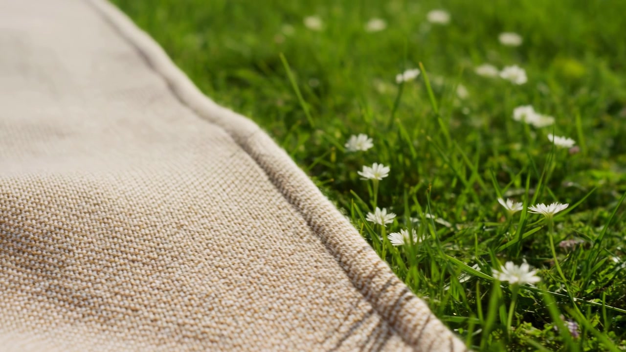 Una vista ravvicinata mostra erba verde con fiori bianchi accanto a un materiale intrecciato beige, con luce diurna delicata, texture fini e una tranquilla disposizione esterna da una prospettiva bassa con toni naturali