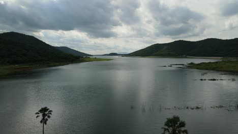 reverse aerial footage over the lake and palm trees also revealing cloudy sky, a lovely horizon, mountains and hills; muak klek, saraburi, thailand