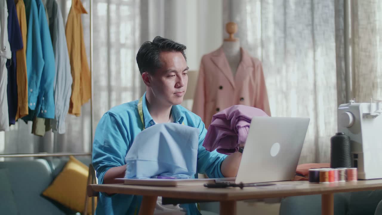 Asian Male Designer With Sewing Machine Holding And Looking At The Fabrics While Designing Clothes In The Studio