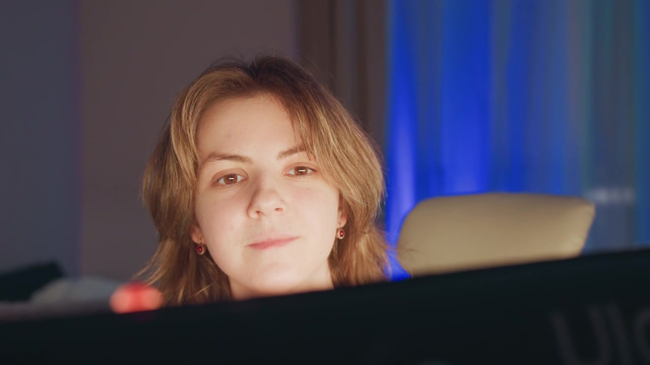 Medium close-up of young woman leaning forward behind monitor edge, looking toward camera with warm attentive expression and subtle smile; screen glow lights face in cozy indoor scene