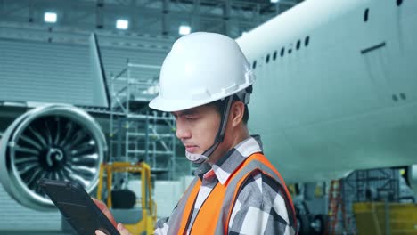 vista lateral cercana de un ingeniero masculino asiático con casco de seguridad trabajando en una tableta mientras está de pie con aviones en el hangar
