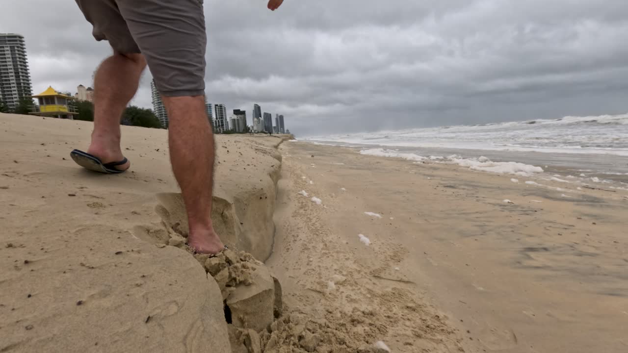 플립플로프를 입은 남자가 침식된 해변 가장자리에 미끄러져 해안 침식과 흐린 하늘 아래의 폭풍의 영향을 강조합니다.