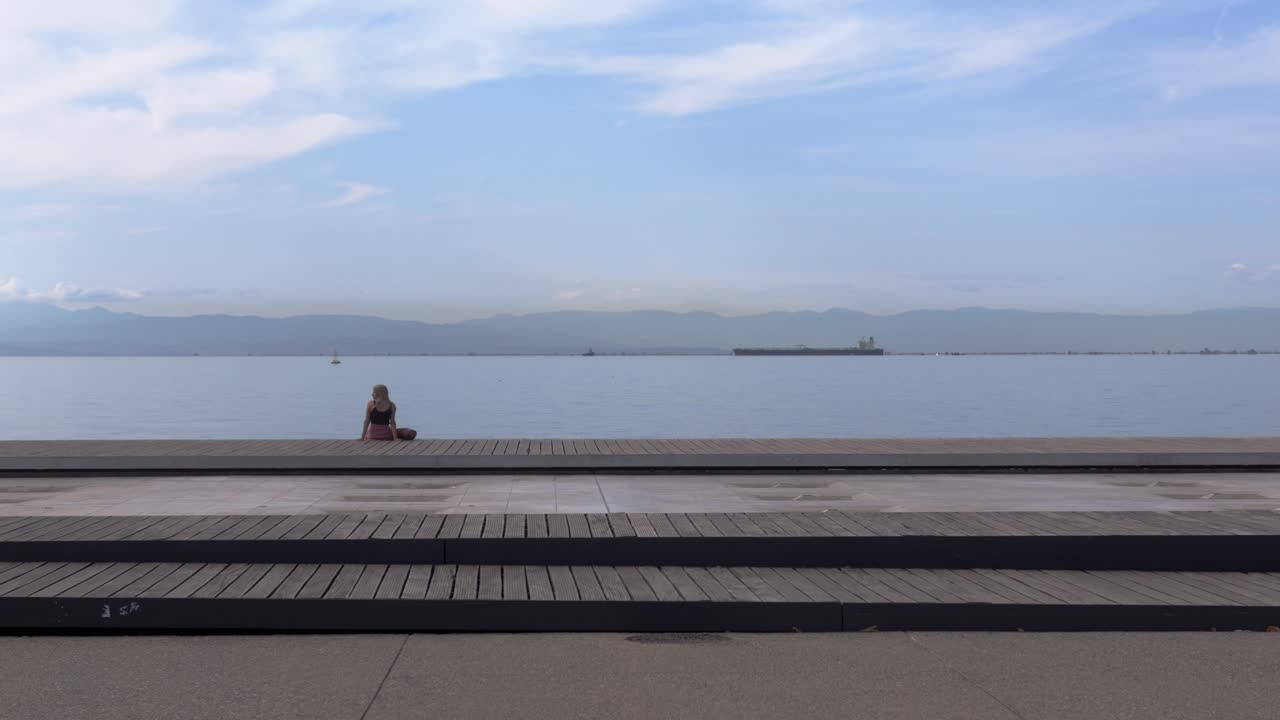Woman sitting on a pier staring at the ocean from the harbor with boats in the background