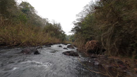 low aerial footage capturing a river winding through the lush jungle, with rocks and vibrant vegetation lining the riverbanks beneath a cloudy sky