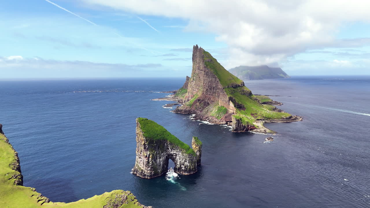 Cinematic aerial view of Drangarnir sea stacks rising dramatically from the Atlantic Ocean near Vágar, Faroe Islands, showcasing rugged cliffs, lush green slopes, and misty Nordic seascape