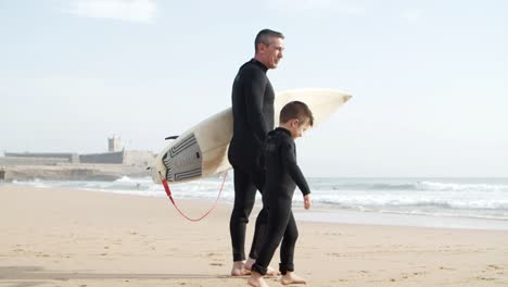 father and son in wetsuits walking on beach