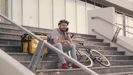 repartidor de comida feliz en un descanso sentado en un escalón hablando por teléfono junto a una mochila térmica