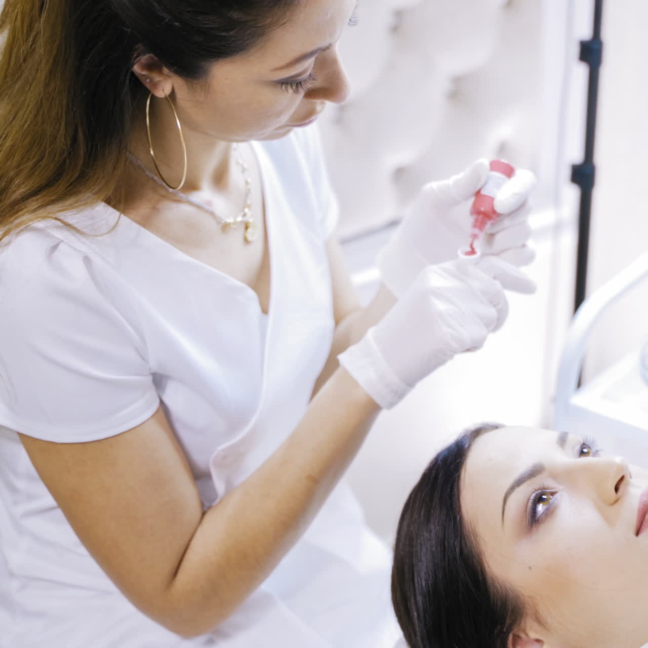 Pretty beautician taking red ink to make permanent make-up in salon. Cosmetologist in protective white gloves preparing equipment to make micropigmentation on lips for a young girl.