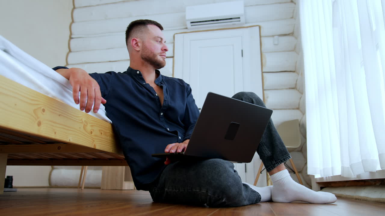 Thoughtful Caucasian man sits on the floor leaning on his bed looking at window. Male holding a laptop on his knee waiting for e-mail message. Work remote.