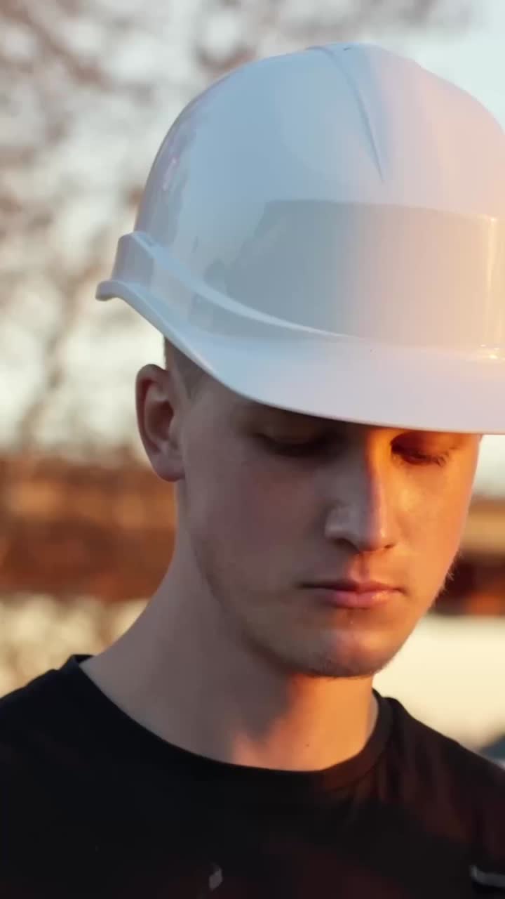 Giovane uomo caucasico con cappello bianco e occhiali di sicurezza trasparenti che sistema i DPI in loco, ritratto di ispettore-ingegnere concentrato che trasmette conformità, sicurezza e responsabilità ambientale, scatto verticale
