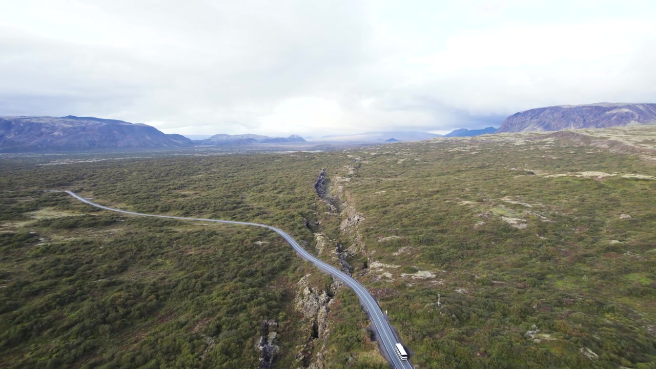 Riprese con drone del paesaggio islandese che mostrano una strada attraverso un terreno aperto, con un autobus che attraversa la scena in una giornata nuvolosa.
