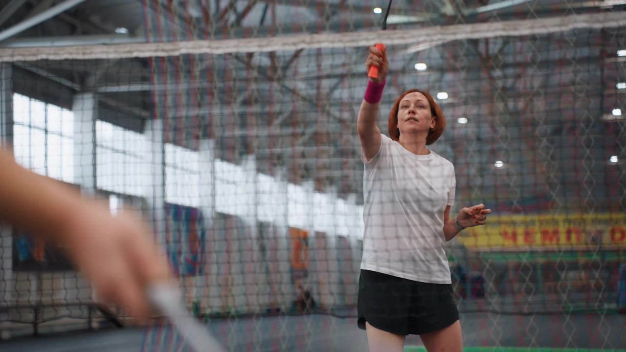 Kaukasisk kvinde server badminton indendørs, øver kontrollerede server og kast, trænerlignende partner giver feedback, net og bannere i baggrunden, træningsøvelse i fitnesscenter, viser koncentreret holdning