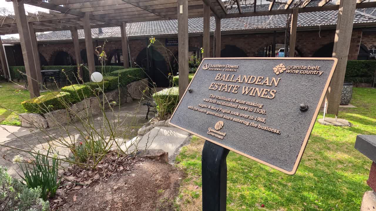 A handheld camera pans across a bronze informational plaque in a landscaped winery courtyard, with bright natural sunlight and lush greenery visible