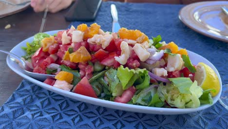 Exprimir-Limones-En-Una-Deliciosa-Y-Saludable-Ensalada-Con-Queso-Feta,-Tomates,-Lechuga,-Cebolla-Y-Naranjas,-Sabroso-Almuerzo,-Toma-De-4k