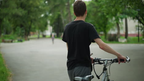 young boy in black top and ash pants walks alongside his bicycle in quiet pathway, while a child approaches him in the distance, the background features softly blurred greenery