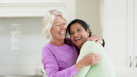 Dos-Mujeres-Mayores-Felices-Y-Diversas-Abrazándose-Y-Sonriendo-En-Una-Cocina-Soleada,-En-Cámara-Lenta