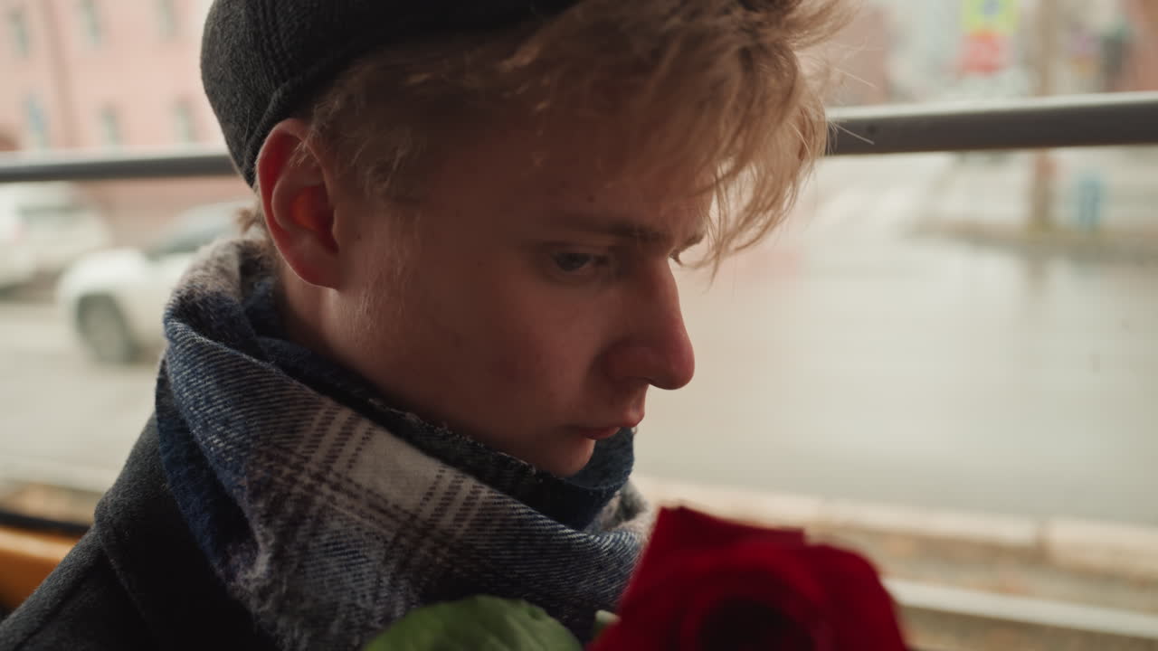 Pensive white male creator on bus holding red rose, wrapped in wool scarf and beanie, watching rainy city through window, cinematic portrait with quiet romantic mood, slow motion commuter atmosphere