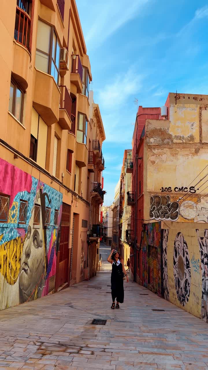 Madrid, Spanyol, 8 Agustus 2025: Jalan kota sempit dengan mural warna-warni dan seorang wanita berjalan. Pemandangan luas lorong beraspal di Spanyol menampilkan seni grafiti di dinding dan seseorang yang berjalan.