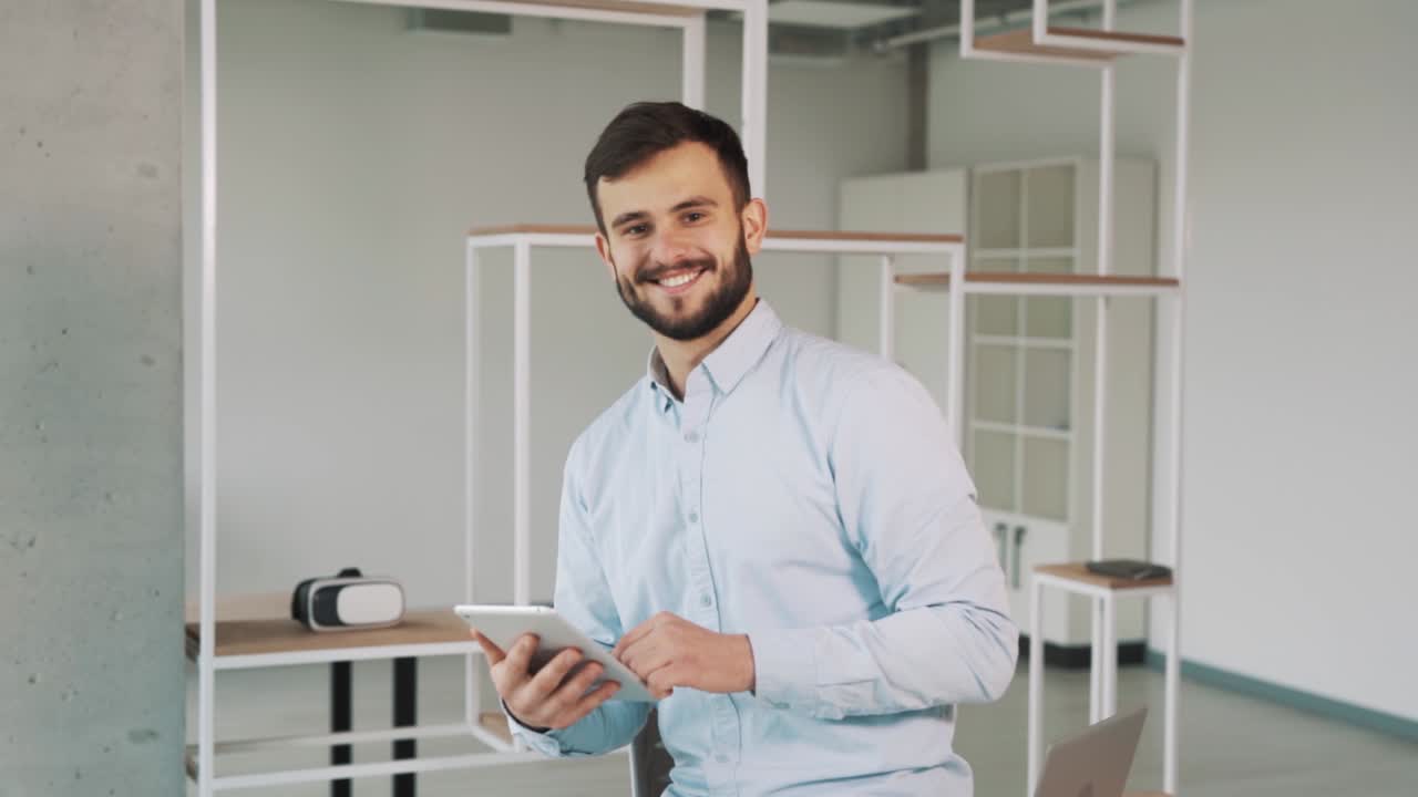 A man in a shirt holding a tablet in his hands is looking at the camera and smiling in a new stylish office