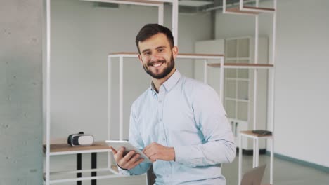 Ein-Mann-Im-Hemd,-Der-Ein-Tablet-In-Der-Hand-Hält,-Blickt-In-Die-Kamera-Und-Lächelt-In-Einem-Neuen,-Stilvollen-Büro