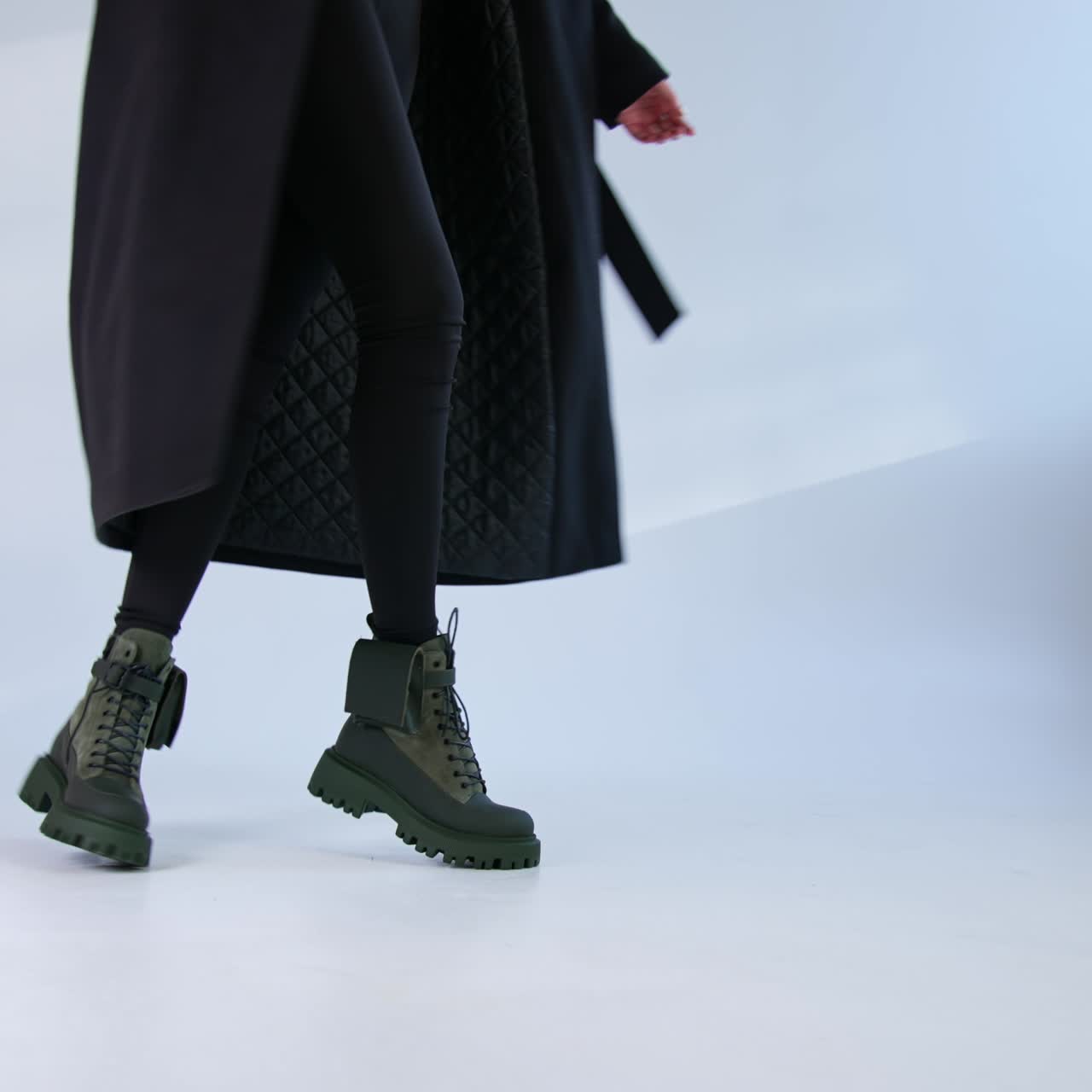 Female model wearing dark leggings and black coat turning in her army boots. Modern stylish boots on the white floor backdrop