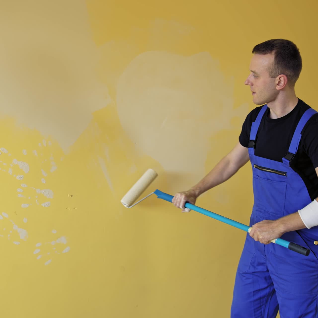 Man is painting walls at home. Young builder in blue uniform renovating walls with a paint roller on a long handle. Painting services