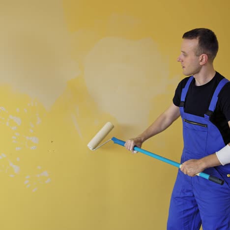 man is painting walls at home. young builder in blue uniform renovating walls with a paint roller on a long handle. painting services