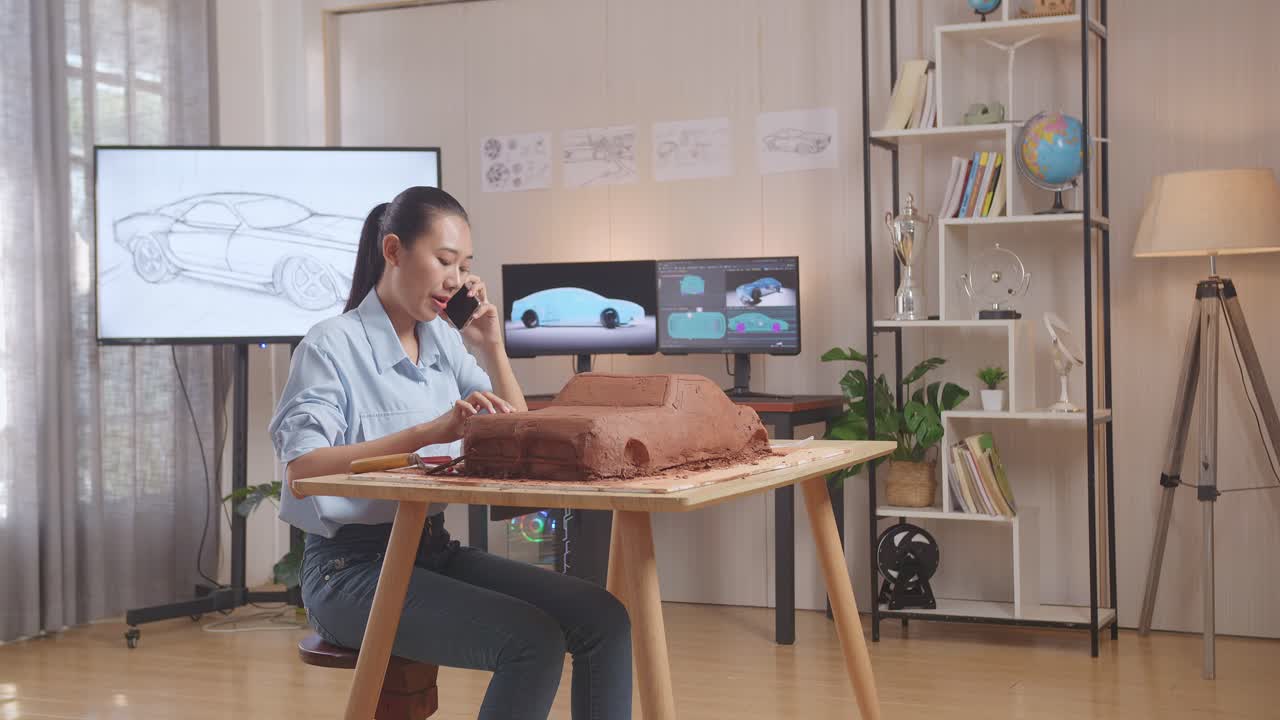 아시아 여성 자동차 디자이너가 스튜디오에서 자동차 점토 조각을 작업하는 동안 스마트폰으로 이야기합니다.