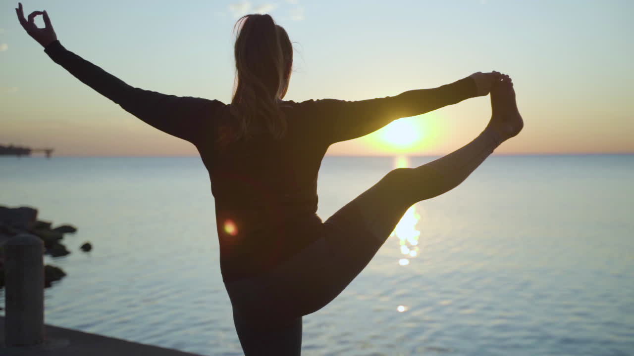 Woman getting into balancing yoga pose by the water at sunrise. Slow Motion