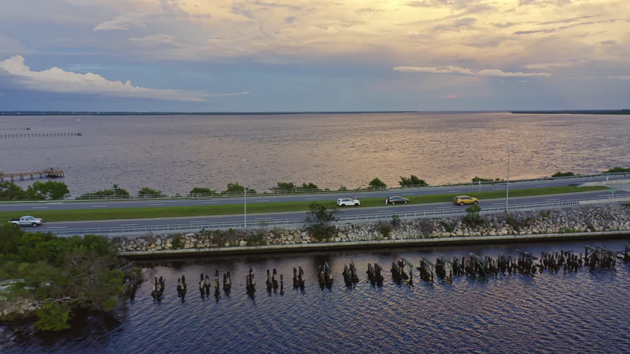 Vehicles travel along a quiet coastal road lined with greenery and rocks as the setting sun casts warm reflections across the still bay beneath a glowing sky