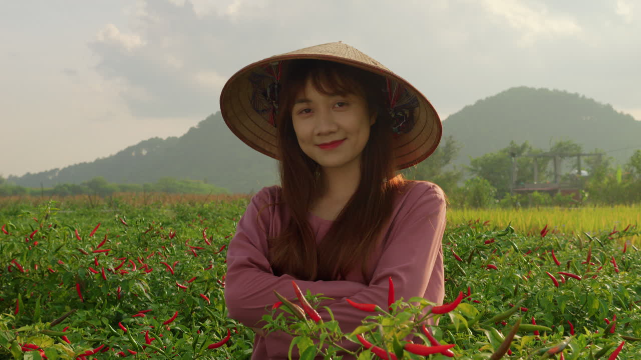 은 뜨거운 칠리 고추 농장에서 해가 지는 동안 카메라 앞에서 미소 짓는  농부 모자를 입은 아시아 베트남 젊은 소녀