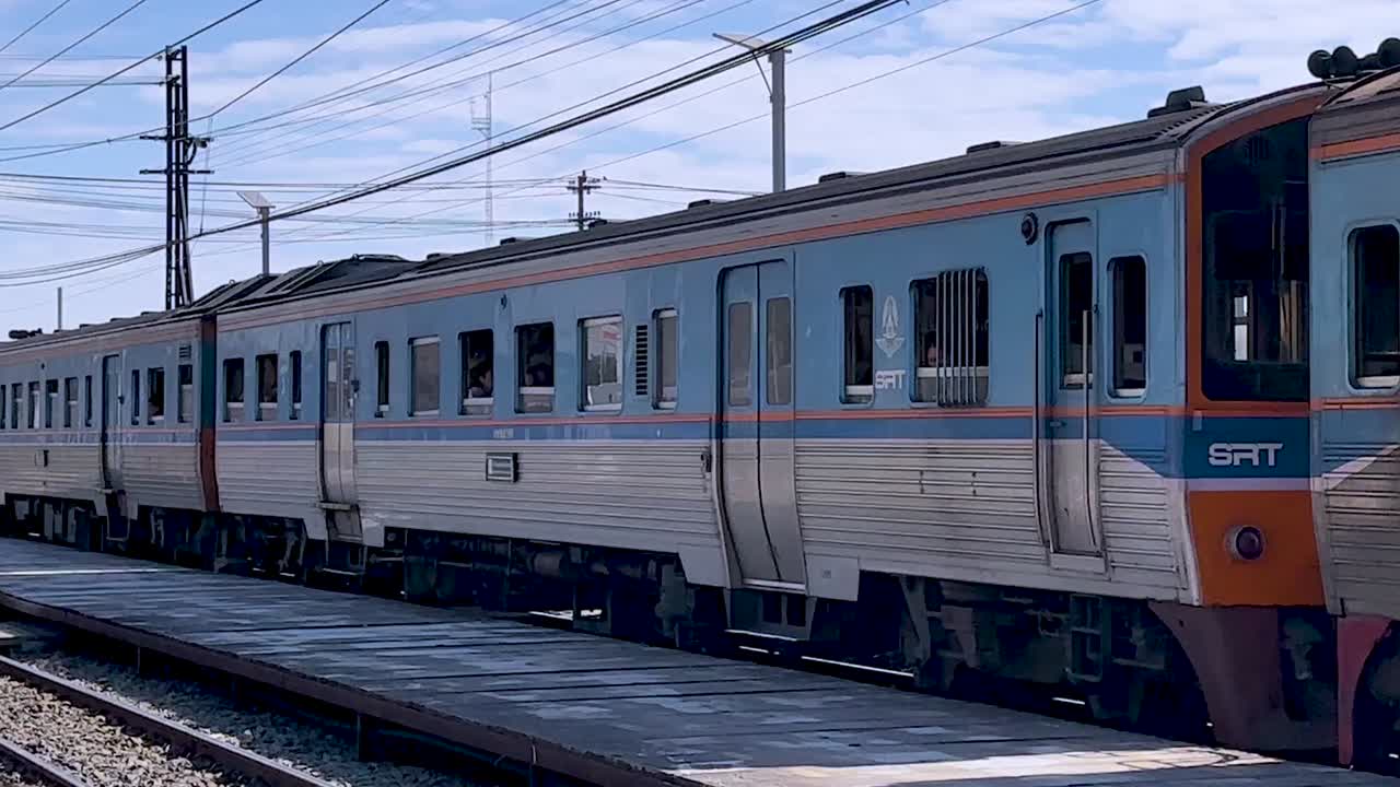 Un treno pendolari blu e argentato passa lentamente davanti a una piattaforma ferroviaria rurale in una giornata di sole.