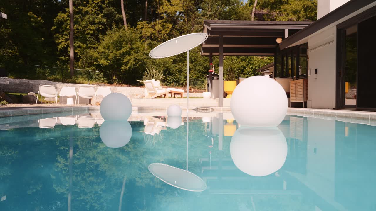 panning shot of a pool outside of a luxury home with three large floating white balls in the pool