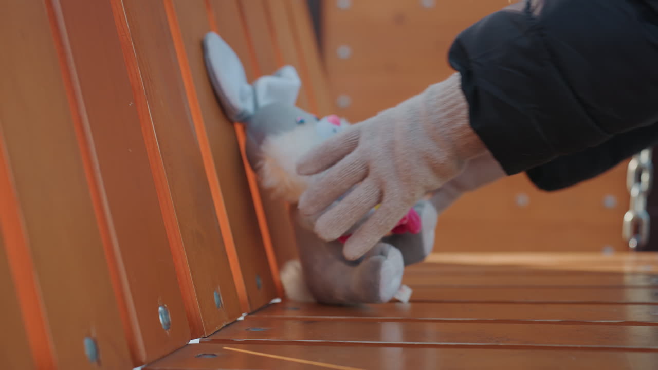 Stuffed bunny on bench, gloved hand removes plush snatcher implied as wintergloved hand takes abandoned toy from wooden park bench near chained swing, deserted playground atmosphere, tension