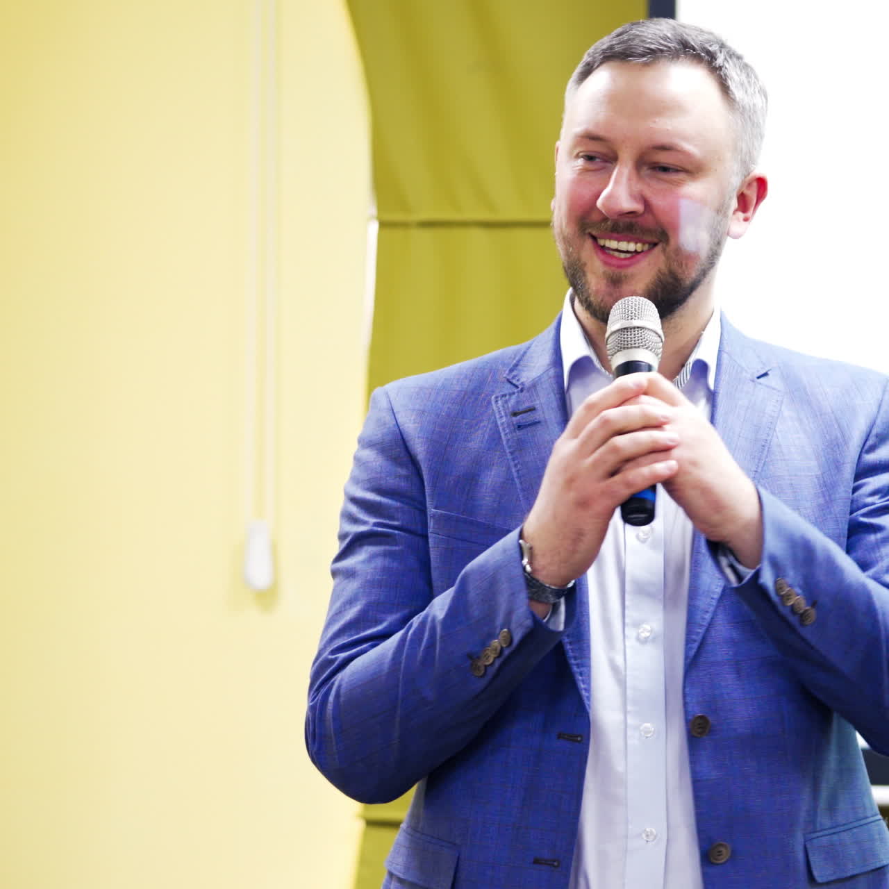 Professional coach in white shirt and blue suit speaking into the microphone in conference hall. Handsome man speaker conducting a lecture with a smile on his face.