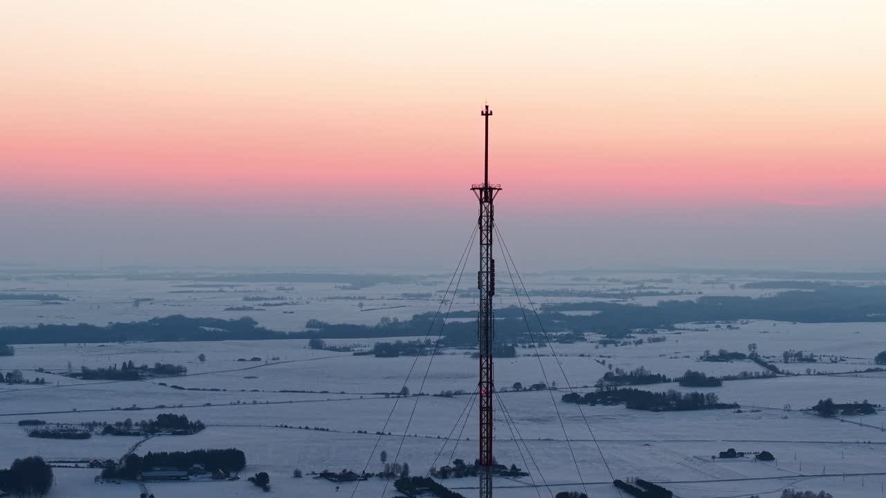 Ripresa con drone che cattura un'alta torre di comunicazione in mezzo a un vasto paesaggio innevato all'alba. Il cielo sfumato di rosa e blu aggiunge un'atmosfera serena ed eterea. Perfetto per scene invernali.