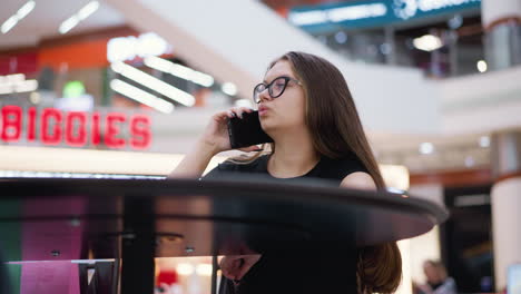 young woman in glasses talking on phone, glancing to left with thoughtful expression, seated in modern setting with blurred background featuring contemporary structures and bright indoor lighting