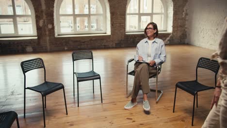 A-girl-psychologist-with-a-bob-haircut-in-a-light-shirt-is-waiting-for-participants-in-group-therapy-who-come-and-sit-in-a