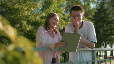 young woman holding tablet and smiling while standing next to man in casual clothing leaning on railing, both enjoying outdoor interaction with blurred greenery in background