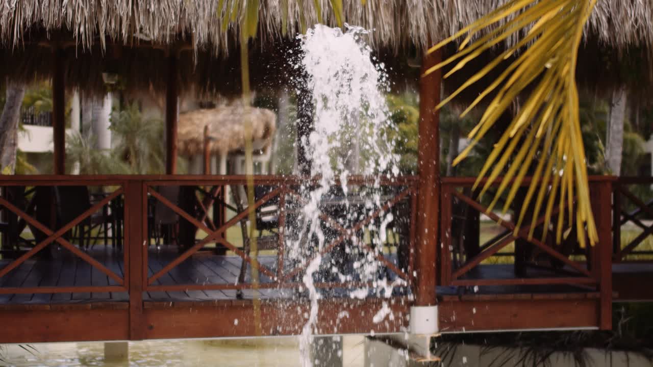 Cinematic slowmo of water fountain next to thatched gazebo over water
