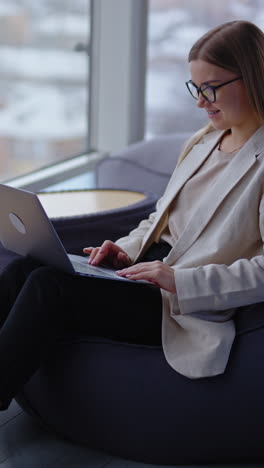 comfortable workplace in fashionable new office premise. lady sitting in a bean bag chair working on laptop. panoramic window backdrop. vertical video