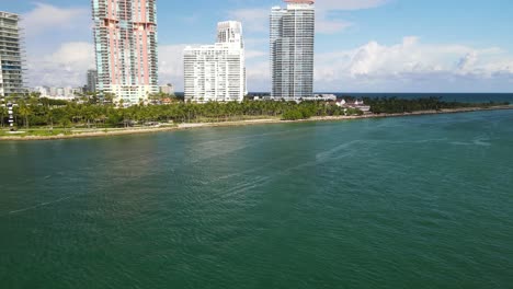 Rascacielos-Costeros-Entre-Palmeras-Tropicales-Con-Agua-De-Mar-De-Marea-Verde-Tropical-Y-Cielos-Azules-Con-Nubes-Suaves